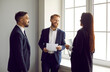 © Studio Romantic - Business colleagues in formal suits have pleasant conversation during break in office. Two men and woman working in same company discuss documents and business projects standing by bright window.