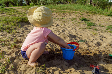 little baby playing with a blue plastic bucket filled with water on a background of sand, a child with bare feet pours water from a bucket learns something new fun childhood in the summer on vacatio