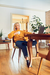 Wall Mural - Cheerful relaxed woman using laptop while at home drinking coffee