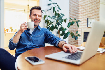 Relaxed smiling man sitting at dinning table at home dinking coffee using laptop
