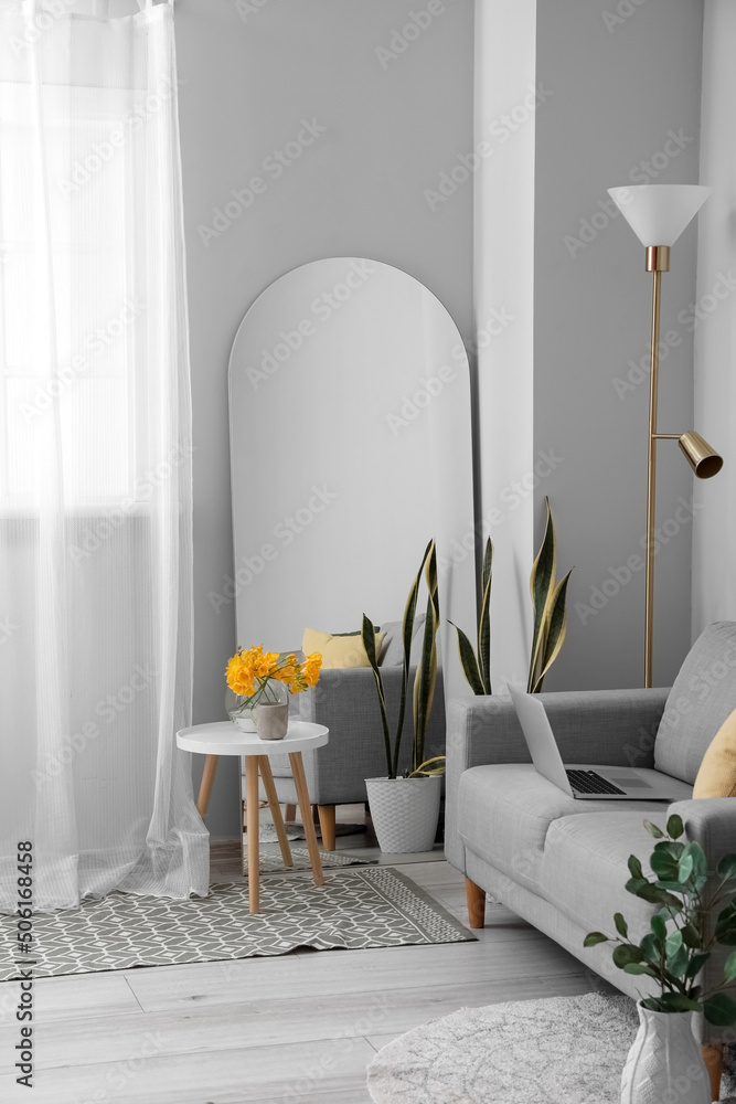 Beautiful flowers on table, big mirror and comfortable sofa in living room interior