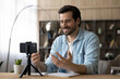 © fizkes - Modern way of teaching. Inspired millennial male school teacher record video lesson at home sit at desk before smartphone on holder talk on web cam. Young man vlogger influencer shoot speech on cell
