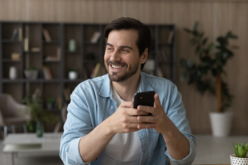 Wall Mural - dreamy and inspired. happy casual young man holding smartphone looking at distance thinking about pl