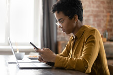African Woman Sit At Desk Holds Smartphone. Distracted From Work On Wireless Computer, Lead Sms Chat With Client, Provide Support Online, Sync Information From Cellphone To Laptop Through App Concept