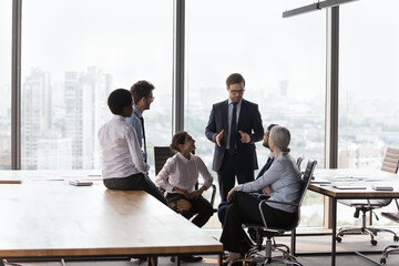 Wall Mural - Millennial multi ethnic team engaged in teamwork at group meeting lead by confident man boss in suit makes speech, tell about corporate goals in office boardroom. Business training, coaching concept