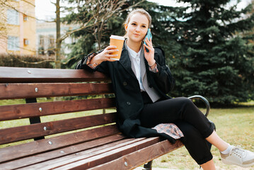 Wall Mural - Pretty smiling young woman calling on smartphone during coffee break while sitting on park bench. Smiling pretty woman wearing stylish coat talking on mobile phone and holding cup of coffee outdoors