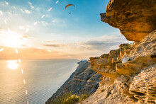 Paraglider Flying From The Rocks Over The Sea Against The Sunset Sky