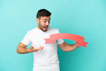 Wall Mural - Young handsome caucasian man isolated on blue background holding an empty placard with surprised expression