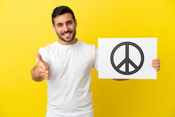 Wall Mural - Young handsome caucasian man isolated on yellow background holding a placard with peace symbol making a deal