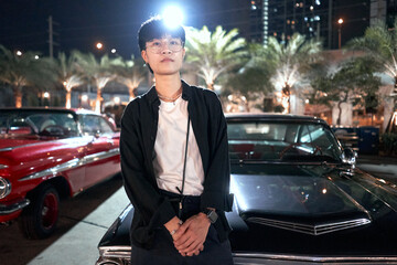 Wall Mural - Serious non-binary person with glasses leaning on a car in an urban night fair