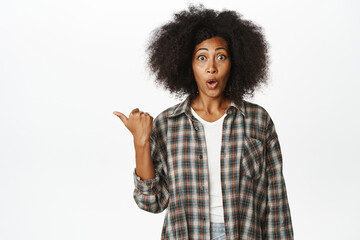 Wall Mural - Surprised african girl points left with amazed gasp and disbelief, asking question about store advertisement, intrigued with promo offer, white background