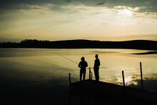 Two Men Silhouette Fishing. Sunset Landscape. Wooden Pier On A Lake. Sunrise Catching Fish With A Rod. Tranquil Water Surface. Fishing Pond Background.