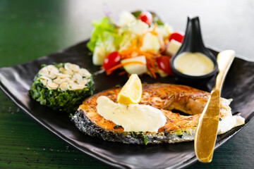 salmon fish steak grill lemon with nut in spinach and vegetable fruits salad with sauce and tomato on black dish and green table with gold or brass knife for seafood and omega 3 in Japanese restaurant