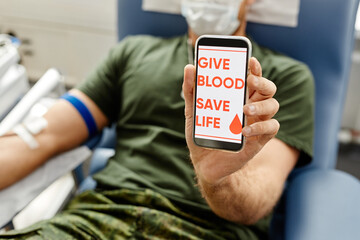 Wall Mural - Closeup of soldier donating blood and holding smartphone to camera with Give blood save life on screen, copy space