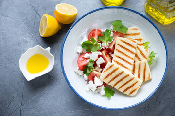 Wall Mural - Plate with grilled halloumi, red tomatoes, feta and parsley, high angle view on a grey concrete background, horizontal shot