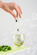 Female hand pours liquid chlorophyll into a glass of water with a dropper. Glass of liquid chlorophyl and fresh herbs on the green table. Gray background, natural light. Concept of superfood, healthy