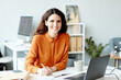 © Seventyfour - Horizontal medium portrait of joyful young adult woman working in office making notes in notebook smiling at camera