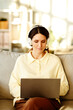 © Seventyfour - Vertical shot of beautiful young adult Caucasian woman wearing casual clothes and headphones sitting on sofa at home working remotely using laptop
