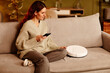© Seventyfour - Modern young adult Caucasian woman wearing casual clothes sitting on sofa at home adjusting settings of robotic vacuum cleaner