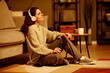 © Seventyfour - Stylish Caucasian woman wearing casual clothes sitting relaxed on floor in living room listening to music in wireless headphones in evening