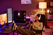 © Seventyfour - Modern Caucasian woman wearing casual clothes and headphones sitting with feet on desk spending evening playing video games with use of controller