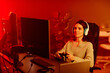 © Seventyfour - Joyful young adult Caucasian woman playing shooter or racing video game using console controller in living room at night, neon red light