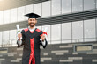 Front view of student with master degree standing, holding two diplomas, smiling. Handsome male in mortarboard and graduate gown graduating from high school. Concept of youth.