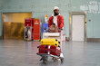 © DimaBerlin - Young African American hipster man pushing luggage trolley while walking after arrival at airport terminal. Trendy Black male tourist rolling a baggage cart in terminal. Trip, journey concept