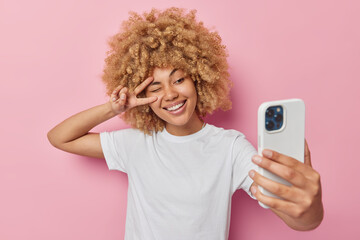 Wall Mural - beautiful young woman with curly blonde hair makes peace gesture over eye poses at smartphone camera