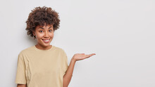 Horizontal Shot Of Pretty Curly Haired Woman Raises Palm Over Blank Space Suggests To Place Promo Text Or Logo Banner There Dressed In Casual T Shirt Isolated Over White Background. Advertisement
