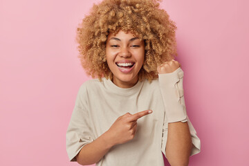 Positive curly haired young European woman points at her arm wears bandage feels happy to overcome challenge wears casual t shirt isolated over pink background. Glad female model cures sprain