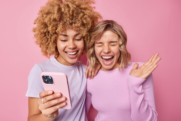 Wall Mural - overjoyed young women laugh happily watch video online hold mobile phone dressed in casual clothes e
