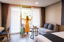 Young Woman Traveler Opening The Curtains And Looking At The View From The Window Of A Hotel Room While On Summer Vacation, Travel Lifestyle Concept