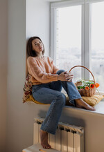 Mature Woman Sitting On Window Sill With Basket Of Tulips At Home