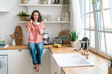 Happy Young Woman Eating Breakfast Sitting On Kitchen Counter At Home