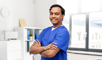 Wall Mural - healthcare, profession and medicine concept - happy smiling doctor or male nurse in blue uniform with crossed arms over medical office at hospital background