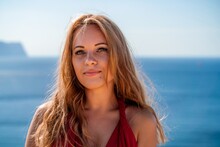 Smiling Young Woman In A Red Dress Looks At The Camera. A Beautiful Tanned Girl Enjoys Her Summer Holidays At The Sea. Portrait Of A Stylish Carefree Woman Laughing At The Ocean.