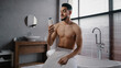 © Yuliia - Arab Indian guy muscular naked sexy unshaven man wears white bath towel on hips sits in bathroom getting ready for bathing washing sniffing shower gel morning refreshment with natural male cosmetics