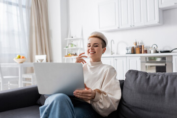 Wall Mural - excited woman with trendy hairstyle laughing near laptop on sofa at home