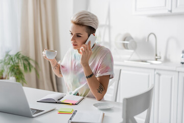 Wall Mural - tattooed woman holding coffee cup during conversation on smartphone near laptop and notebooks