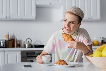Wall Mural - cheerful woman holding coffee cup and eating tasty croissant near smartphone with blank screen