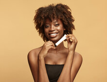 Smiling African American Woman With A Clean And Healthy Skin Holding Slim Cream Tube Over Beige Background.