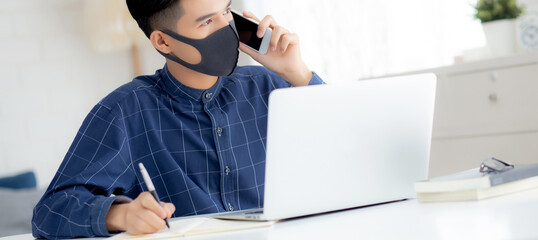 Wall Mural - Young asian business man in face mask talking smartphone and work on laptop computer for protect covid-19, businessman quarantine work from home and writing notebook, social distancing and stay home.