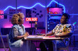 © AnnaStills - Young African American man and woman sitting at table in front of each other recording podcast or broadcast in home studio