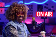 © AnnaStills - Horizontal medium close-up of joyful young African American woman wearing fashionable outfit sitting in broadcasting studio smiling at camera
