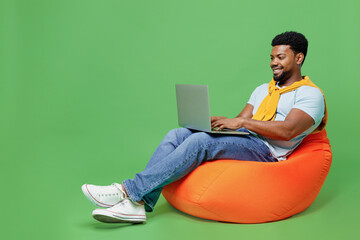 Wall Mural - Full body young man of African American ethnicity 20s in blue t-shirt sit in bag chair hold use work on laptop pc computer isolated on plain green background studio portrait. People lifestyle concept.