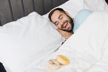 Wall Mural - Young surprised satisfied amazed man wear casual blue t-shirt look at donuts breakfast lying in bed rest relax spend time in bedroom lounge home in own room house wake up dream. Real estate concept.