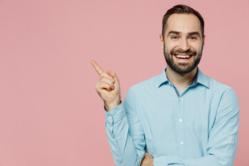 Young smiling fun caucasian man 20s wear classic blue shirt point index finger aside on workspace area mock up isolated on plain pastel light pink background studio portrait. People lifestyle concept.