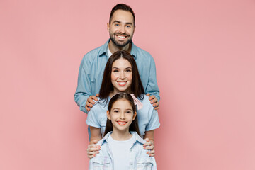 Wall Mural - Young smiling cheerful happy parents mom dad with child kid daughter teen girl in blue clothes stand behind each other look camera isolated on plain pastel light pink background. Family day concept.