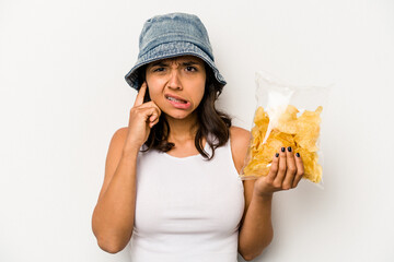 Wall Mural - Young hispanic woman holding a bag of chips isolated on white background covering ears with hands.